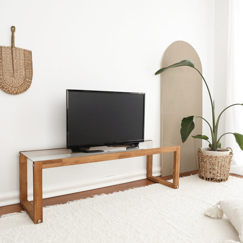 Mobile basso porta tv in stile moderno con gambe in legno massello e ripiano in vetro temperato trasparente, disponibile in finitura noce opaco e rovere chiaro opaco. Dimensioni cm 130x40x40h